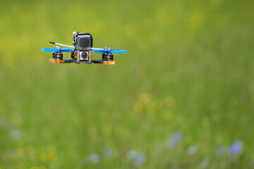 Small racing copter also drone with blue propellers and black battery over a green meadow with colorful flowers. Front view. Depth of field.