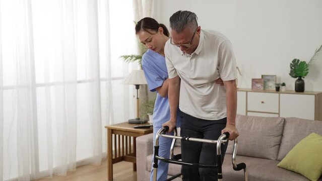 slow motion of asian senior stroke patient undergoing rehab exercise with a walker at home. the woman nursing aide assists him during home visit