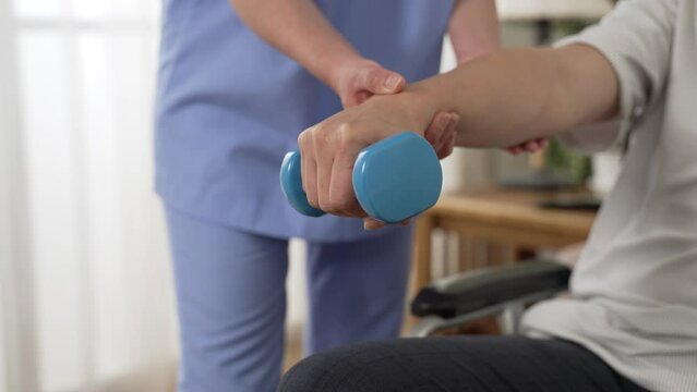 slow motion with closeup hand of wheelchair user holding lifting dumbbell with nurse’s support at background. home health care and fitness for the disabled concept