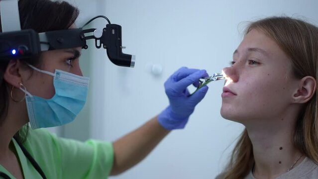 Close-up side view positive otolaryngologist using ent headlight examining nasal canal of patient in hospital. Professional Caucasian woman with nasal forceps checking nose of teenage girl