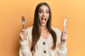 Beautiful brunette young woman holding fork and knife ready to eat celebrating crazy and amazed for success with open eyes screaming excited. - Powered by Adobe