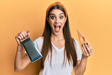 Beautiful brunette young woman holding grater and cheese celebrating crazy and amazed for success with open eyes screaming excited.
