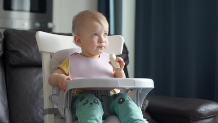 Cute kid sitting in booster seat with feeding tray fixed on top of dining chair, one year old baby sitting in front of tv. High quality 4k footage