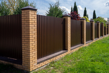 Brown metal fence with brick posts against sky. Outer corner of fence. High wall encloses private area. Security concept