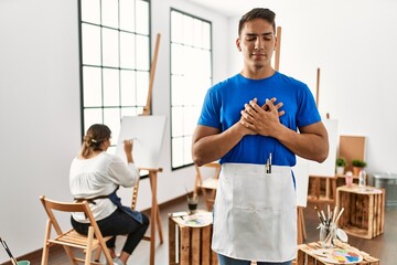 Young hispanic couple at art studio smiling with hands on chest with closed eyes and grateful gesture on face. health concept.