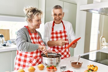 Senior caucasian couple smiling happy baking sweets using touchpad at the kitchen.