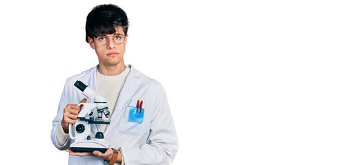 Handsome hipster young man wearing lab coat holding microscope relaxed with serious expression on...