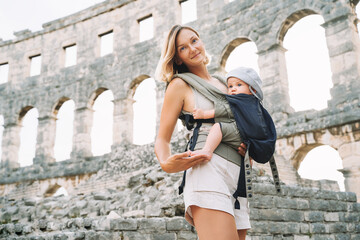 Tourist woman with child in Roman Amphitheater Arena like as Coliseum - famous travel destination in Pula, Croatia. Beautiful mother and baby in carrier on vacation in Europe. Family Active Travel.