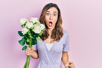 Young hispanic girl holding flowers scared and amazed with open mouth for surprise, disbelief face