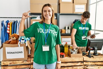Young blonde girl wearing volunteer t shirt at donation stand smiling and confident gesturing with hand doing small size sign with fingers looking and the camera. measure concept.