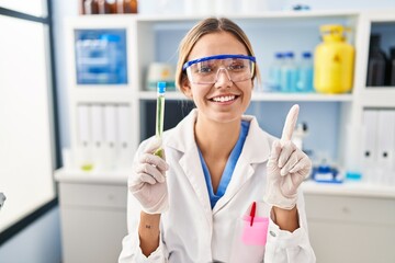 Young blonde woman working at scientist laboratory holding sample smiling with an idea or question pointing finger with happy face, number one