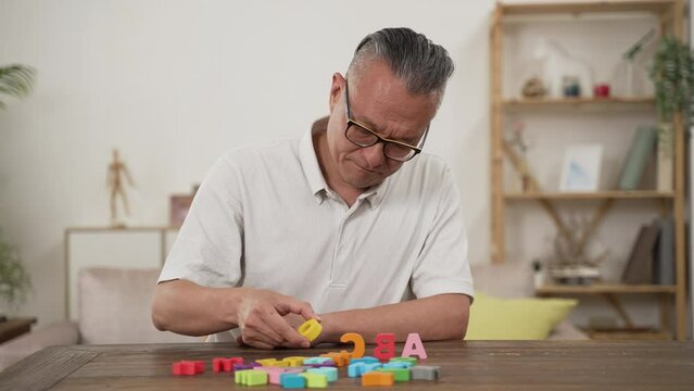 asian Japanese senior male Alzheimer&rsquo;s patient practicing rehab treatment with color letter bricks at home. he compares two blocks and puts them in order on table