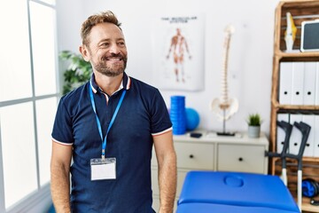 Middle age physiotherapist man working at pain recovery clinic looking away to side with smile on face, natural expression. laughing confident.