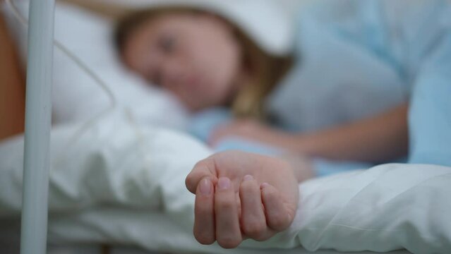 Close-up slim female hand on hospital bed indoors with blurred teenage girl lying at background. Unrecognizable sick Caucasian patient undergoing treatment with eye dropper in clinic