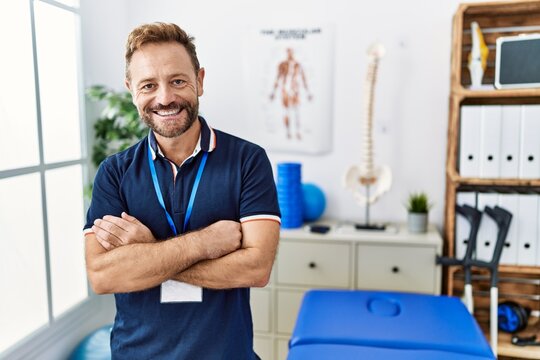 Middle age physiotherapist man working at pain recovery clinic happy face smiling with crossed arms looking at the camera. positive person.