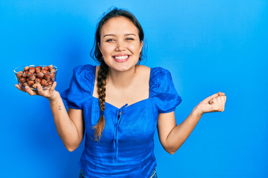 Young hispanic girl holding bowl of chestnuts screaming proud, celebrating victory and success very excited with raised arm