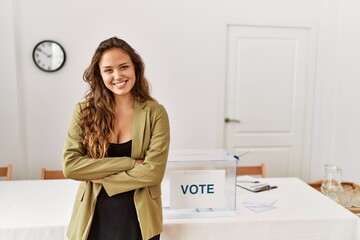 Beautiful hispanic woman standing at political campaign room happy face smiling with crossed arms...