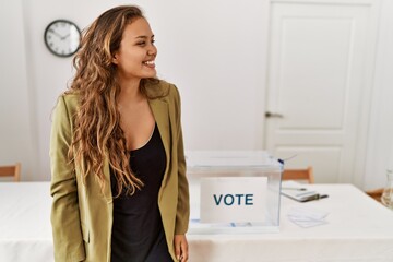 Beautiful hispanic woman standing at political campaign room looking away to side with smile on...