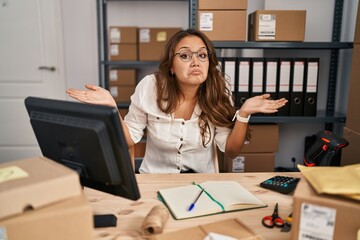 Young hispanic woman working at small business ecommerce clueless and confused expression with arms and hands raised. doubt concept.