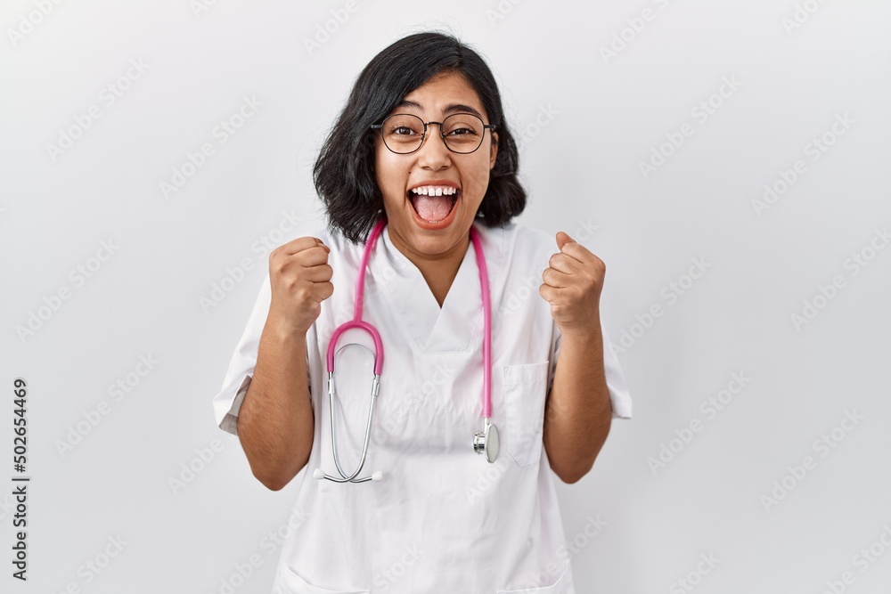 Wall mural Young hispanic doctor woman wearing stethoscope over isolated background celebrating surprised and amazed for success with arms raised and open eyes. winner concept.