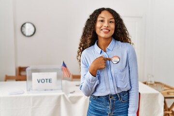 Young latin woman smiling confident pointing with finger to i voted badge at electoral college - obrazy, fototapety, plakaty