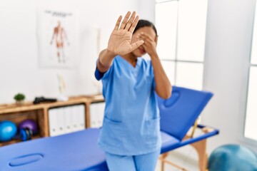 Beautiful hispanic physiotherapist woman at pain recovery clinic covering eyes with hands and doing stop gesture with sad and fear expression. embarrassed and negative concept.