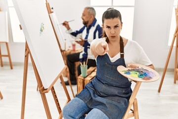 Young artist woman at art studio pointing with finger to the camera and to you, confident gesture looking serious