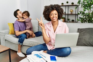 Mother of interracial family working using computer laptop at home smiling looking to the camera showing fingers doing victory sign. number two.