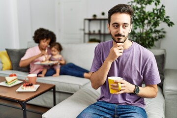 Hispanic father of interracial family drinking a cup coffee with hand on chin thinking about question, pensive expression. smiling and thoughtful face. doubt concept.