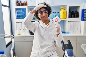 Hispanic man with curly hair working at scientist laboratory holding toxic banner stressed and frustrated with hand on head, surprised and angry face