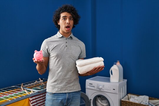 Hispanic man with curly hair holding clean towels and piggy bank afraid and shocked with surprise and amazed expression, fear and excited face.