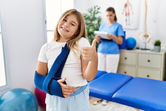 Blonde little girl wearing arm on sling at rehabilitation clinic smiling happy and positive, thumb up doing excellent and approval sign