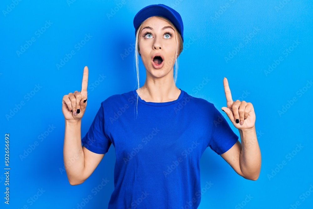 Sticker young caucasian woman wearing courier uniform wearing cap amazed and surprised looking up and pointi