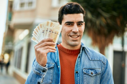 Young hispanic man smiling happy holding denmark krone banknotes at the city.