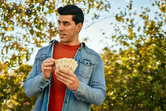 Young hispanic man smiling happy holding denmark krone banknotes at the city.