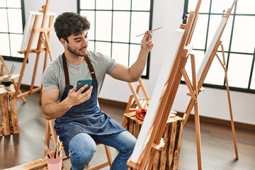 Young hispanic artist man smiling happy painting and using smartphone at art studio.