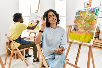 Young hispanic woman at art studio smiling looking to the camera showing fingers doing victory...