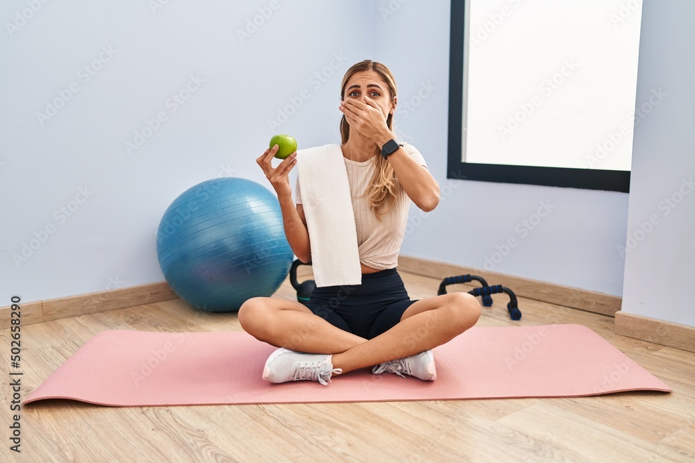 Poster Young blonde woman wearing sportswear and towel holding healthy apple shocked covering mouth with hands for mistake. secret concept.