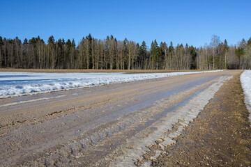 Straight dirt road with mud and melting ice crust among snow-covered farmlands. Hard sunlight on a...
