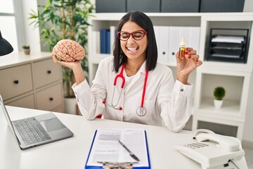 Young hispanic doctor woman holding brain and cbd oil sticking tongue out happy with funny...