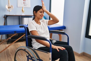 Young hispanic woman sitting on wheelchair at physiotherapy clinic surprised with hand on head for...