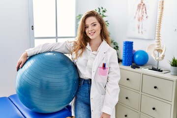 Fototapeta premium Young caucasian woman working at rehabilitation clinic holding pilates ball looking positive and happy standing and smiling with a confident smile showing teeth