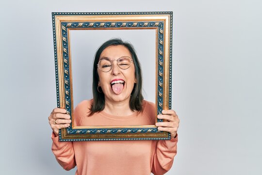 Middle age hispanic woman holding empty frame sticking tongue out happy with funny expression.
