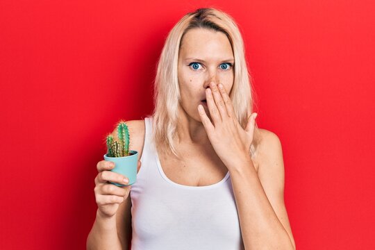 Beautiful caucasian blonde woman holding small cactus pot covering mouth with hand, shocked and afraid for mistake. surprised expression