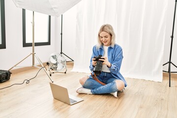 Beautiful blonde woman working as professional photographer at photography studio sitting on the floor checking photos on computer laptop