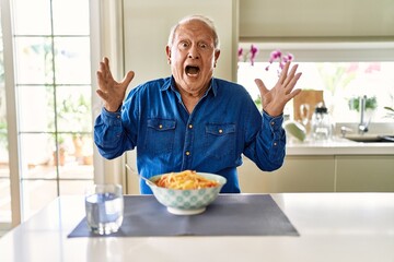 Senior man with grey hair eating pasta spaghetti at home celebrating crazy and amazed for success with arms raised and open eyes screaming excited. winner concept