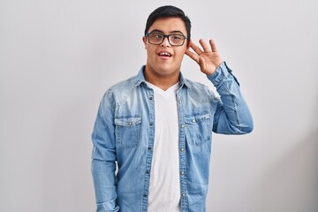 Young hispanic man with down syndrome wearing casual denim jacket over white background smiling with hand over ear listening and hearing to rumor or gossip. deafness concept.