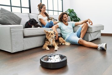 Man and woman couple sitting on sofa with dogs cleaning floor using vacuum robot at home