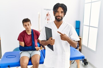 Obraz premium Young hispanic man working at pain recovery clinic with a man with broken arm pointing with hand finger to the side showing advertisement, serious and calm face