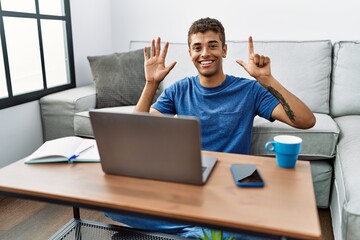 Young handsome hispanic man using laptop sitting on the floor showing and pointing up with fingers number seven while smiling confident and happy.